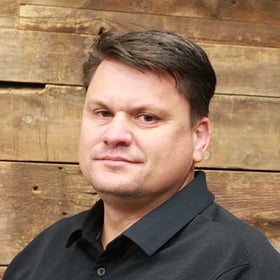 Headshot of David Huff wearing a button-down shirt, smiling in front of a blurred wood background.
