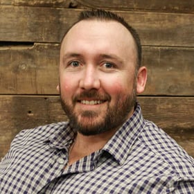 Headshot of Jarid Scarafiotti wearing a button-down shirt, smiling in front of a blurred wood background.