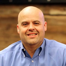 Headshot of Jerry Beighey wearing a button-down shirt, smiling in front of a blurred wood background.