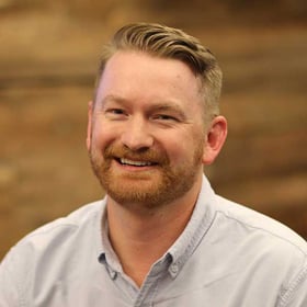 Headshot of Ryan Windham wearing a button-down shirt, smiling in front of a blurred wood background.