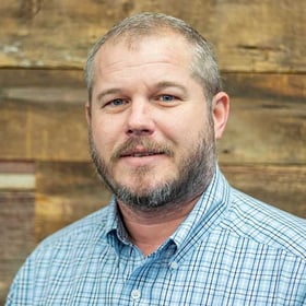 Headshot of Shane Cranford wearing a button-down shirt, smiling in front of a blurred wood background.