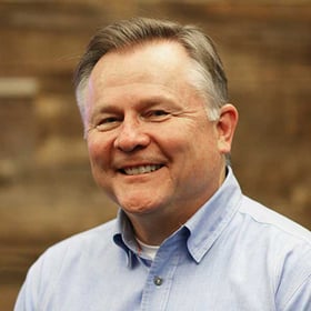 Headshot of Tim Cummings wearing a button-down shirt, smiling in front of a blurred wood background.