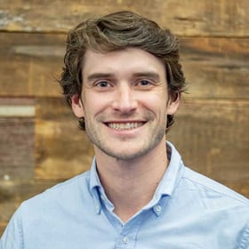 Headshot of Zach Cunningham wearing a button-down shirt, smiling in front of a blurred wood background.