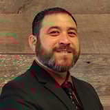 Headshot of Brian Taniguchi wearing a button-down shirt with a tie and black blazer, smiling in front of a wood background.