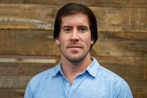 Headshot of Brandon Barajas wearing a button-down shirt, smiling in front of a blurred wood background.