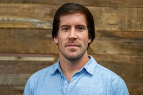 Headshot of Brandon Barajas wearing a button-down shirt, smiling in front of a blurred wood background.