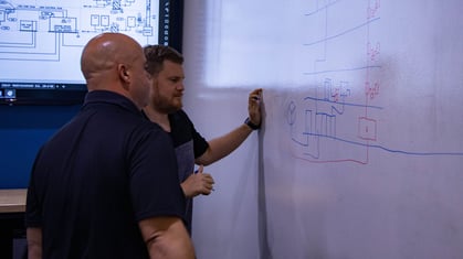 Two men collaborating at a whiteboard with technical diagrams drawn in red and blue marker.