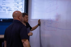 two men writing on a whiteboard with a one-line diagram on a screen behind them