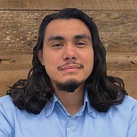 Headshot of Miguel Longoria wearing a button-down shirt, smiling in front of a blurred wood background.