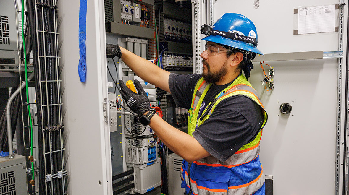 A man in a safety vest is inspecting and working on an electrical panel