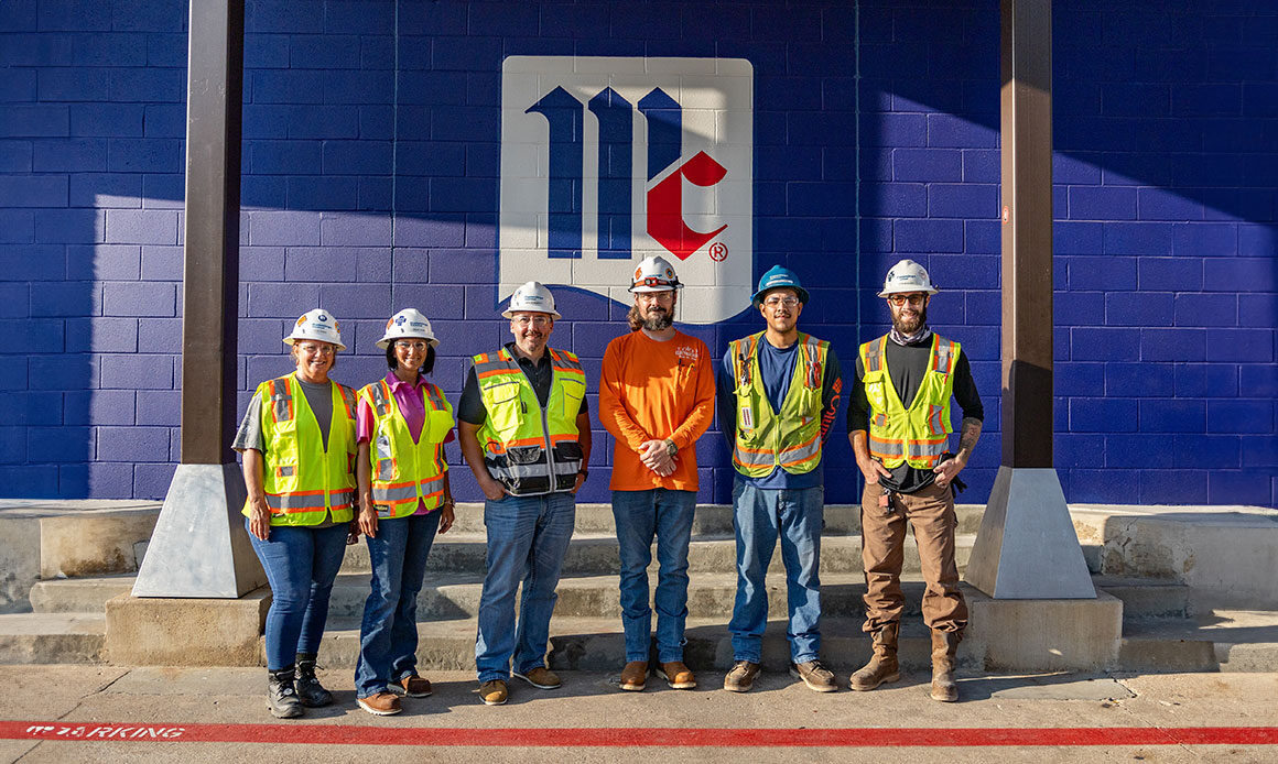 A group of six electrician workers wearing high-visibility vests and hard hats standing in front of a blue wall with the McCormick & Company logo