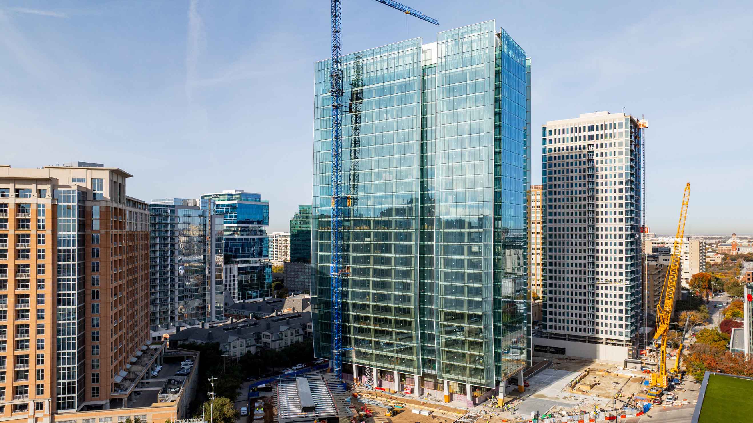 Modern glass office tower under construction in a dense downtown setting with cranes surrounded by high-rises