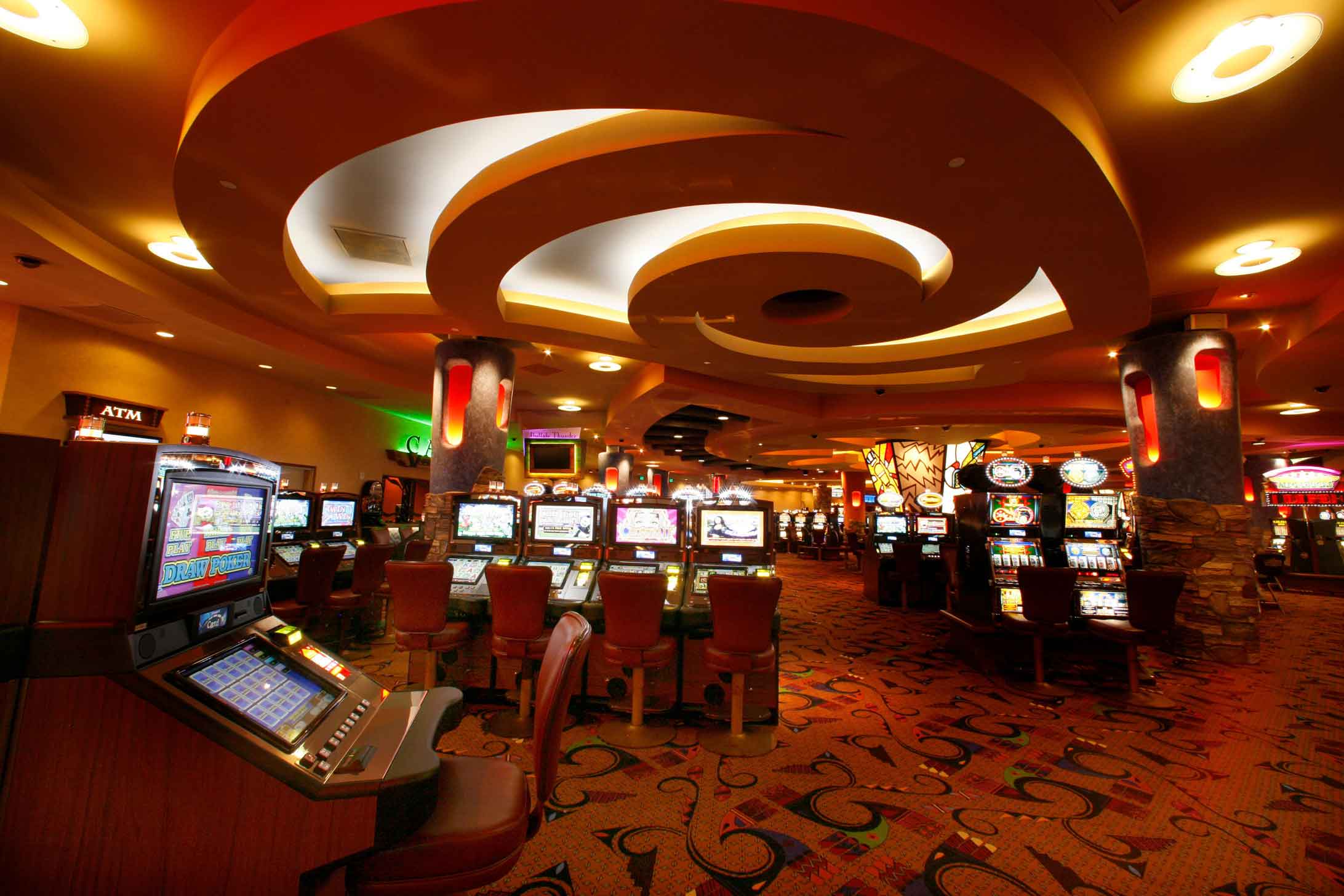 Casino gaming floor illuminated by ambient ceiling lighting and filled with rows of slot machines and patterned carpet