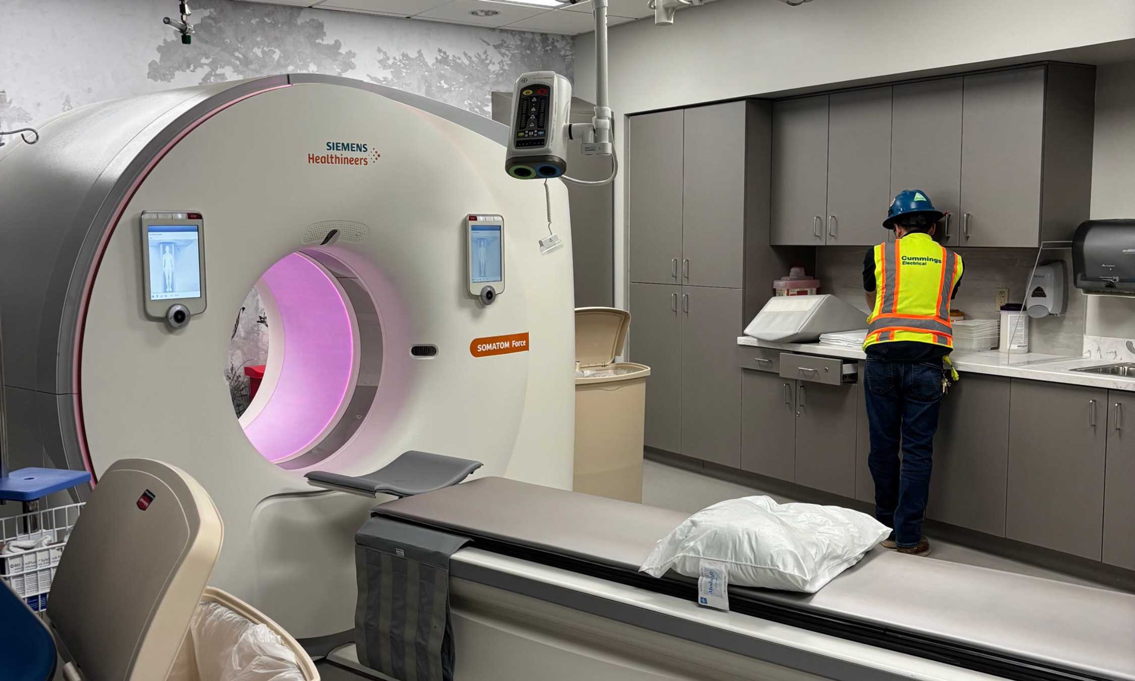 Healthcare construction technician in PPE working beside a CT scanner during installation in a medical imaging suite