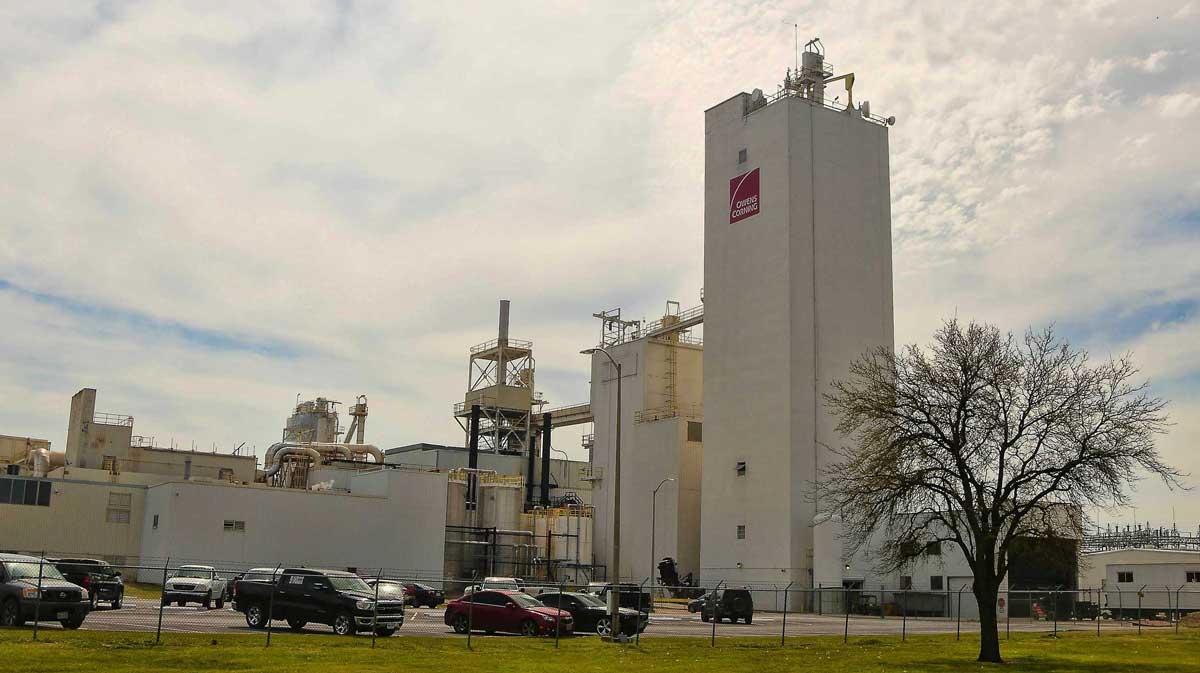 Ground-level view of the Owens Corning manufacturing plant with tall processing towers, support buildings, and a fenced perimeter with parking