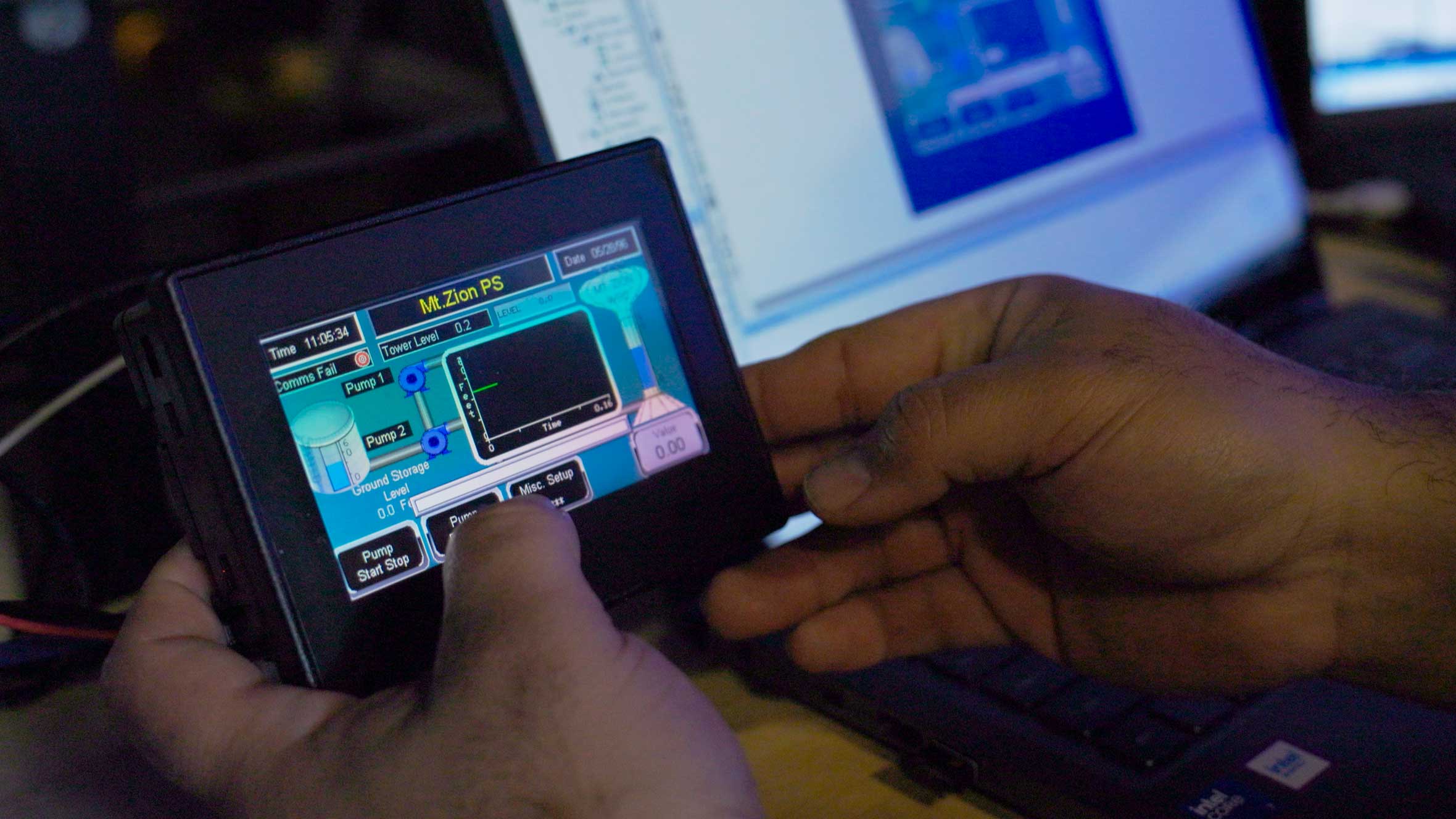Hands holding a small screen with scada system-of-water-facility-with-laptop-in-background