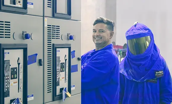 two men in arc gear next to electrical panels and one smiling at the camera