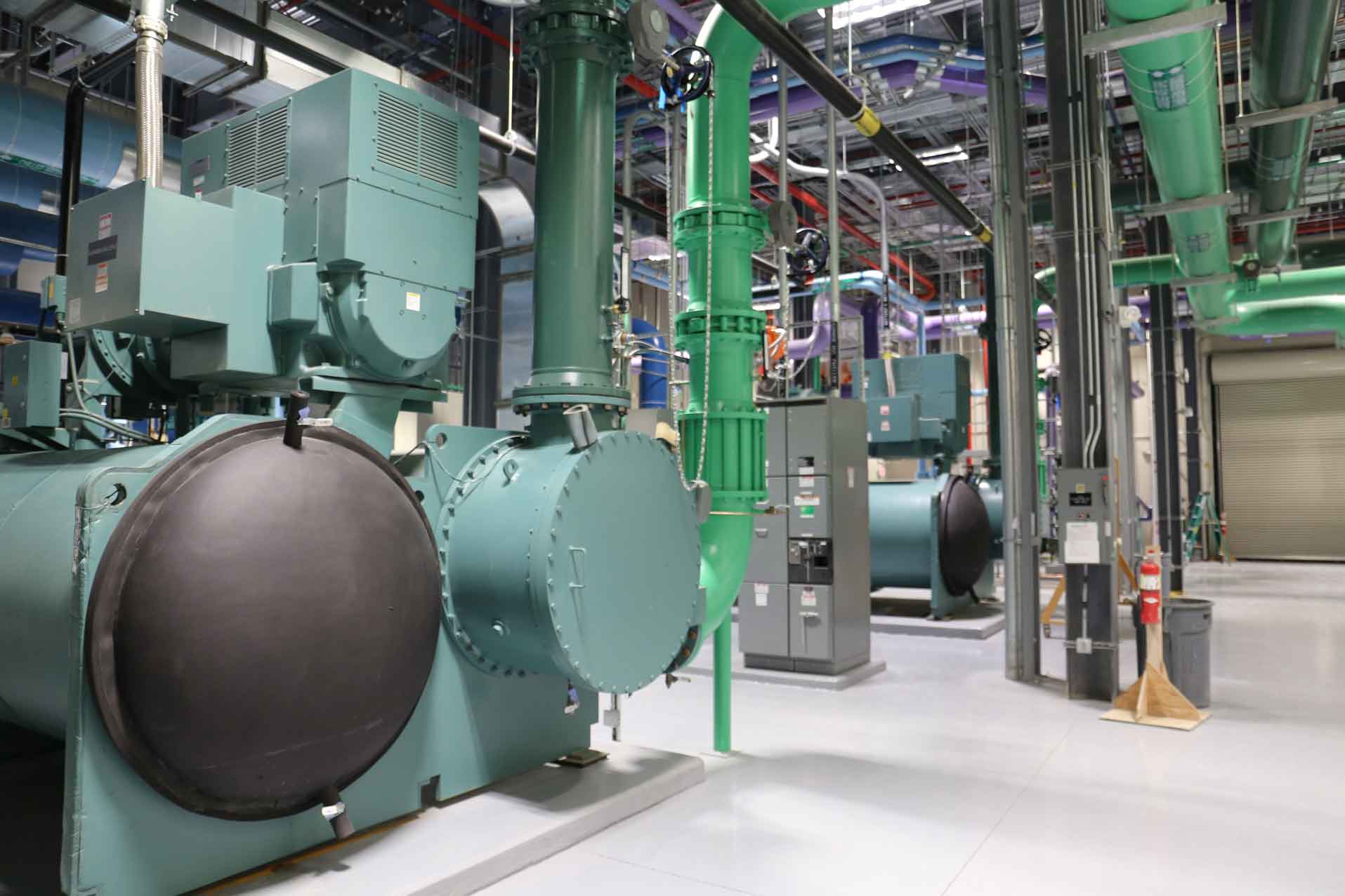 Mechanical room at a children’s hospital with large green chillers, overhead piping, and electrical panels.