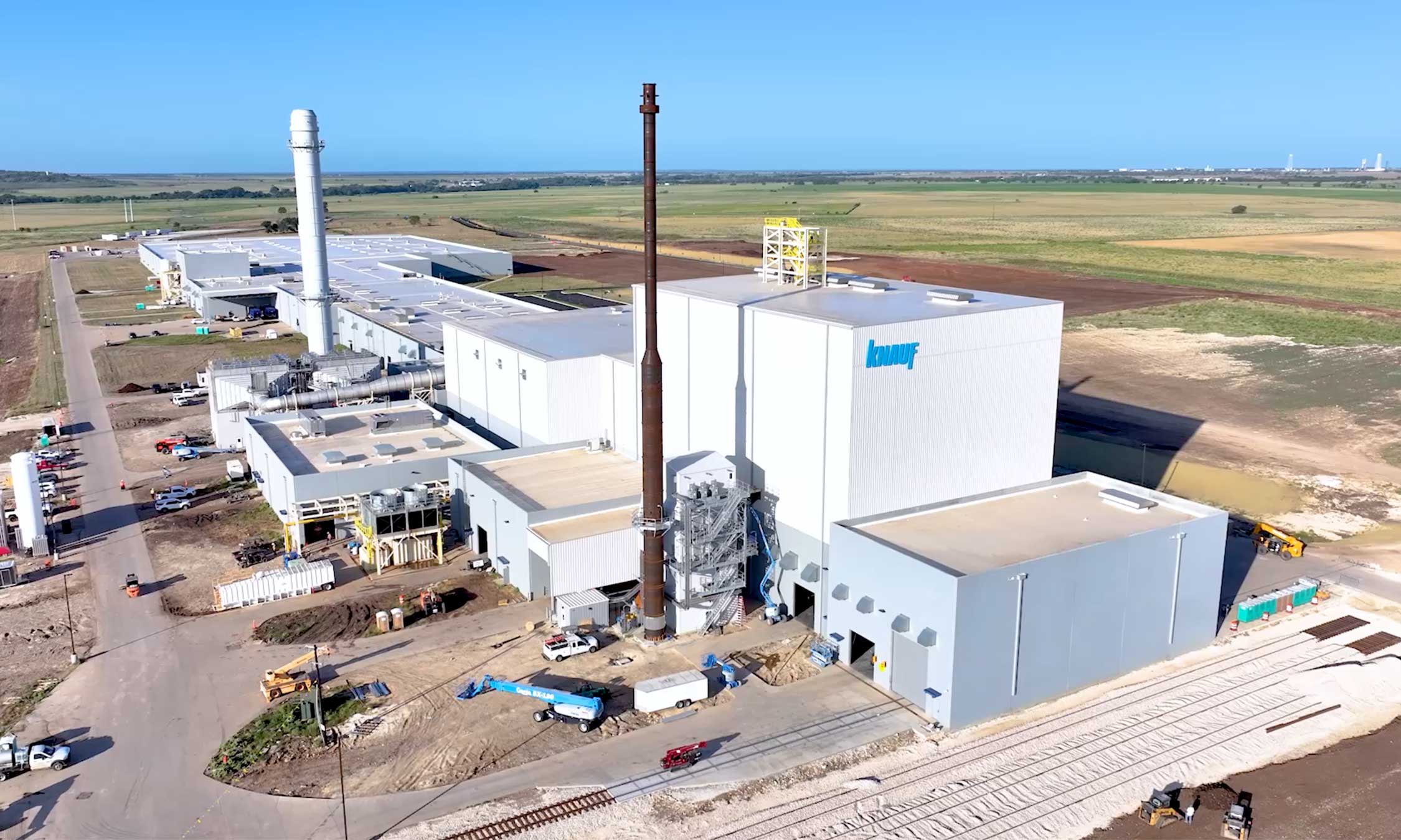 Aerial view of the Knauf insulation manufacturing facility with vertical silos, smokestacks, and active site development