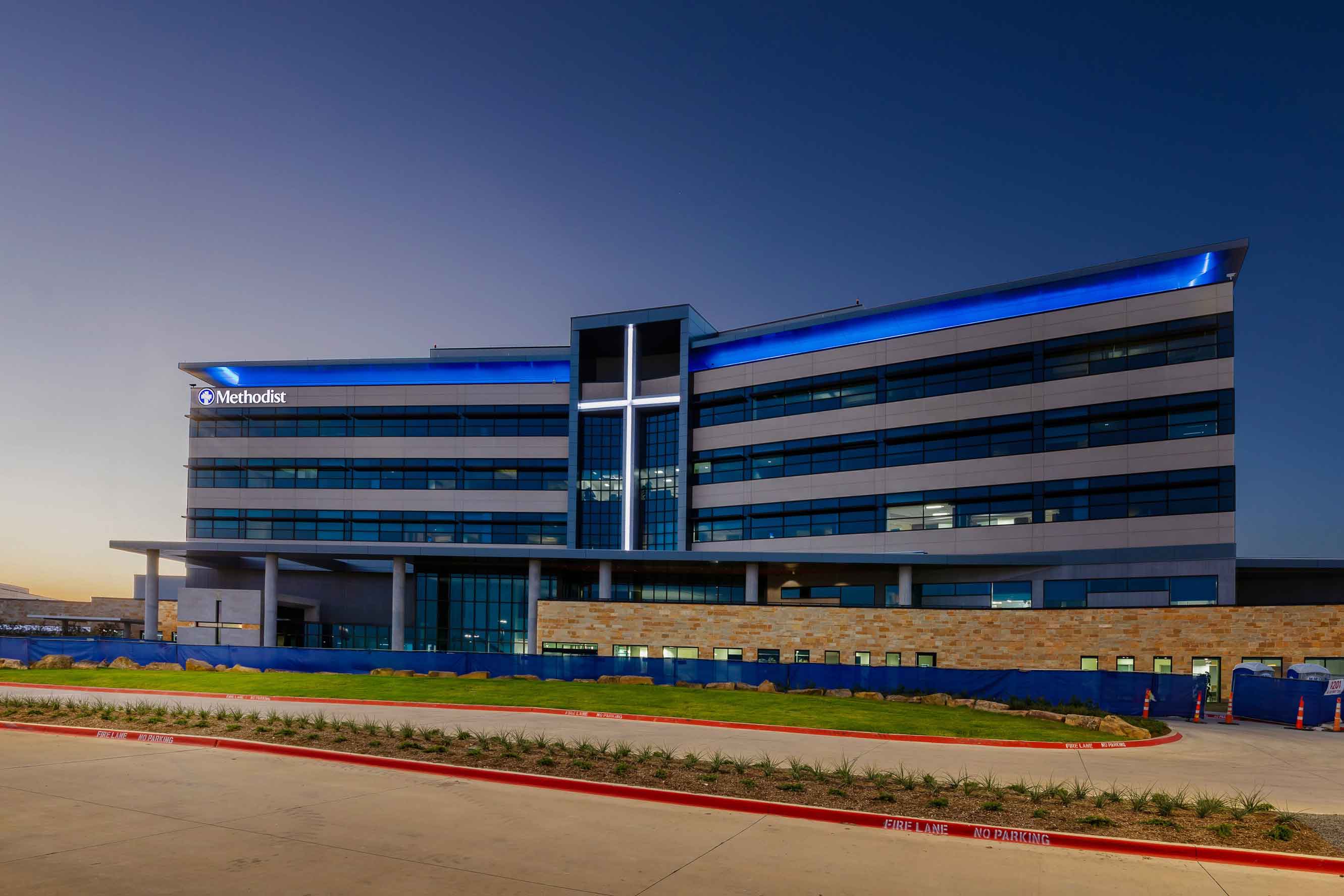 Exterior view of a modern Methodist hospital building with blue lighting on the roof and a large cross illuminated at the front