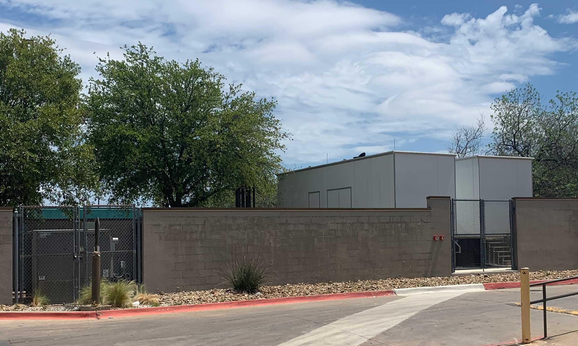 Fenced-in exterior electrical yard behind a masonry wall with visible utility enclosure and service gate