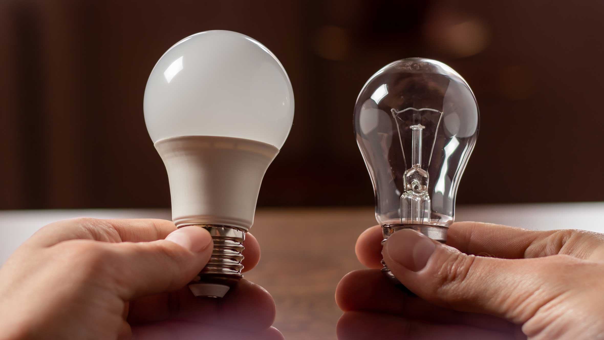 Two hands with one holding an incandescent light bulb and an LED bulb in the other