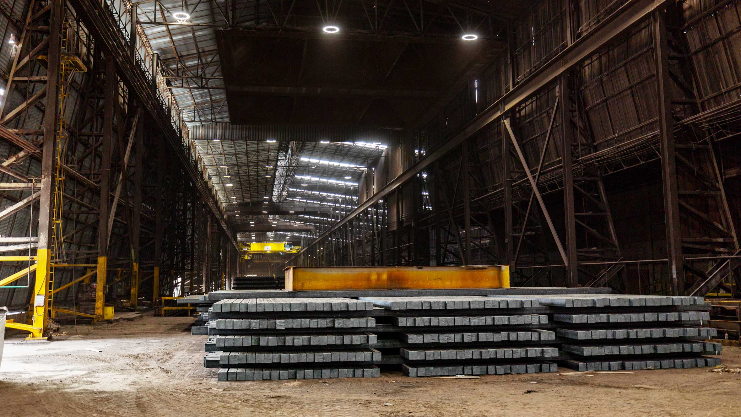 Interior of a heavy industrial facility with stacked steel billets and structural steel framing under a high bay roof